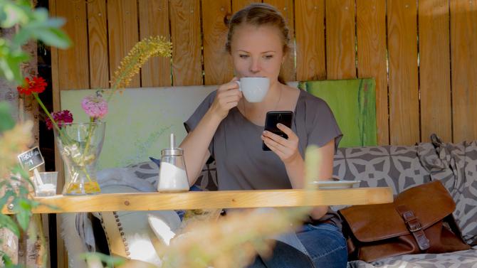 Junge Frau sitzt auf einem Sofa mit einer Kaffeetasse und schaut auf ihr Samrtphone